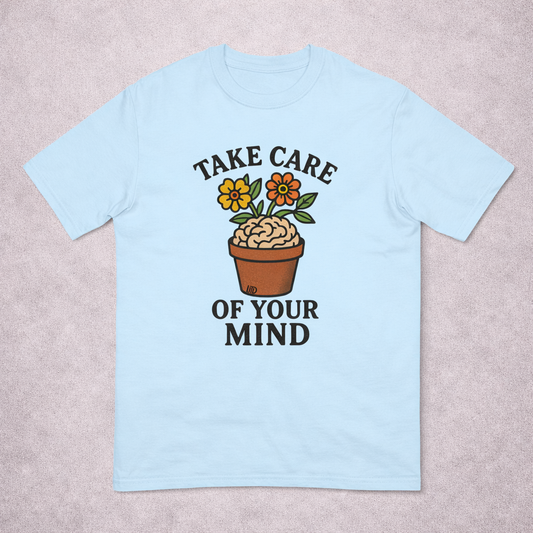 Light blue t-shirt with a brain in a pot and flowers, featuring the text 'Take care of your mind', on a beige background.