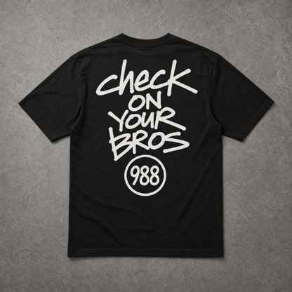 Black t-shirt with white text 'check on your bros 988' on a gray textured background